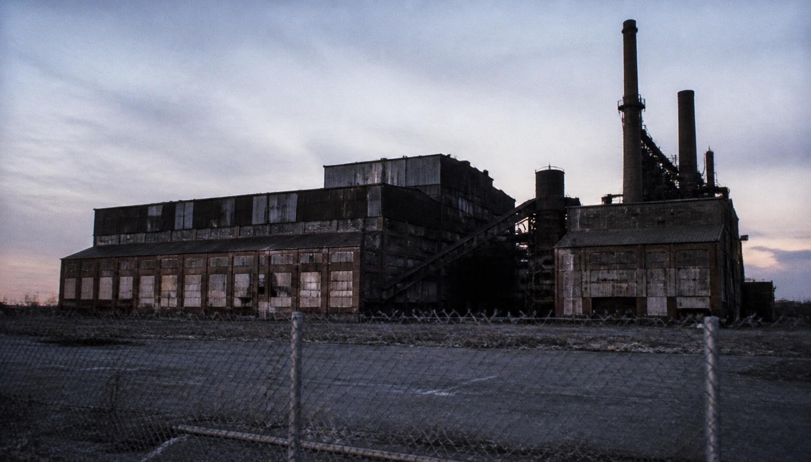 A shuttered American factory at dusk behind a chain link fence