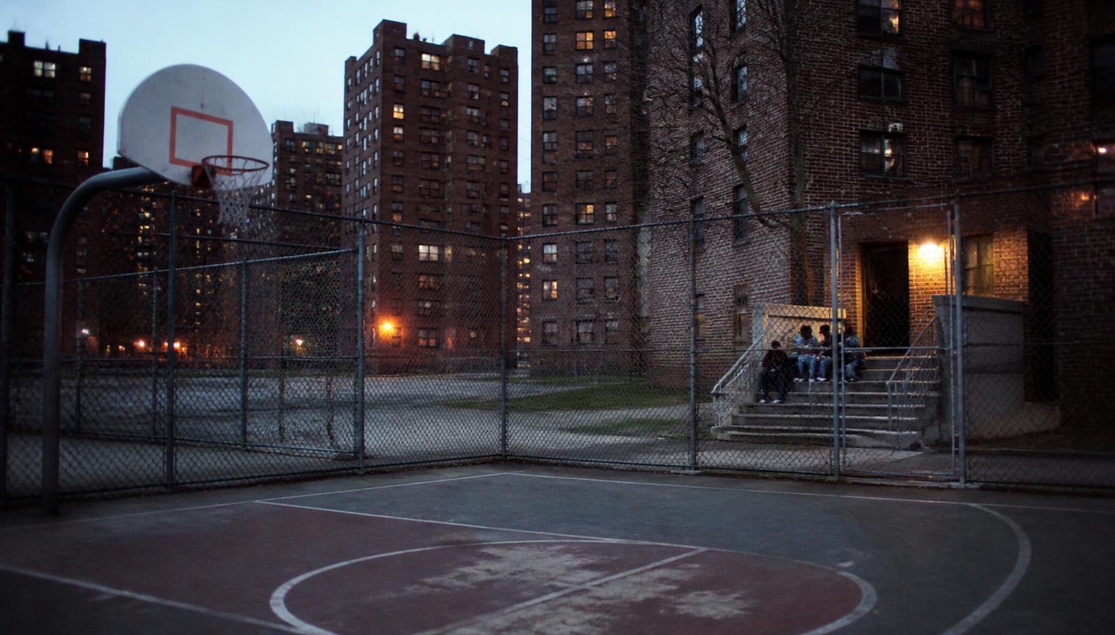 Public housing towers at dusk behind an empty neighbourhood basketball court